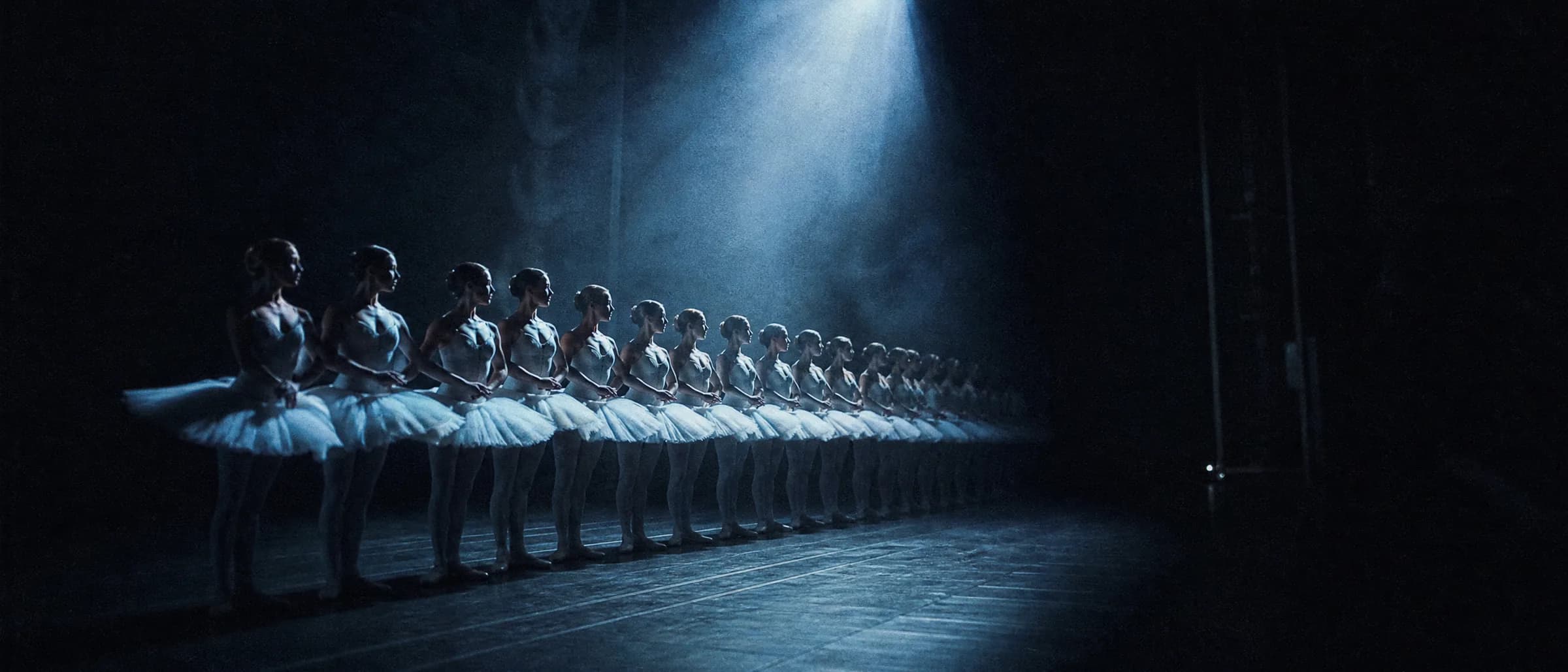 Le corps de ballet aligné en diagonale sous un unique faisceau bleu, le fond de scène perdu dans le noir