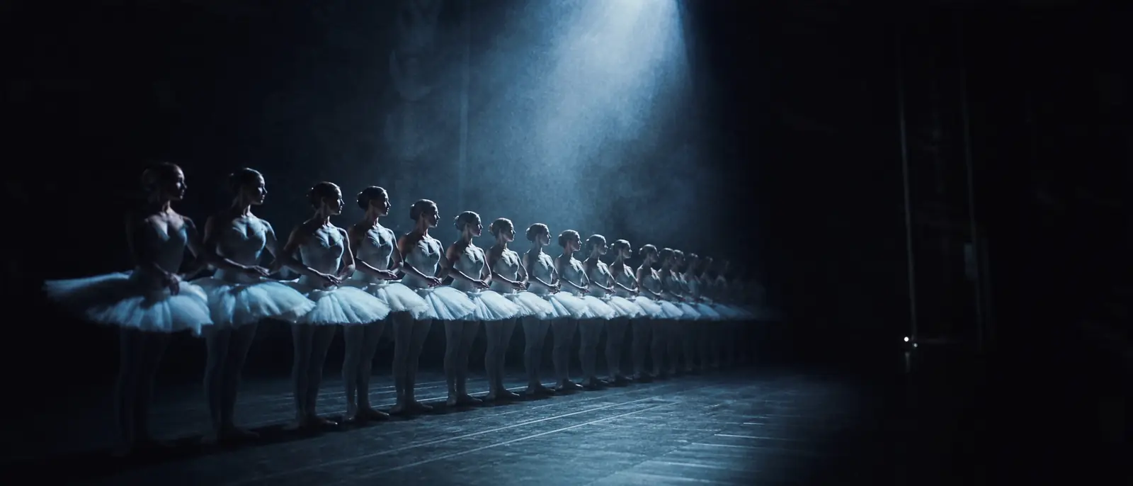 Le corps de ballet aligné en diagonale sous un unique faisceau bleu, le fond de scène perdu dans le noir